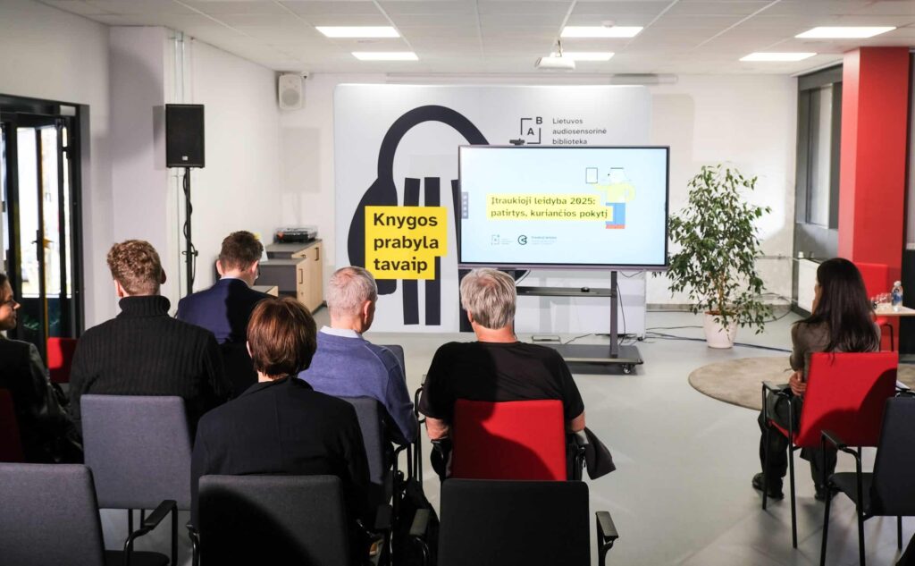 Renginio akimirka: Lietuvos audiosensorinės bibliotekos renginių salė, kurioje matyti iš nugaros sėdintys žmonės, priešais juos – TV ekranas su užrašu: Įtraukioji leidyba 2025: patirtys, kuriančios pokytį. Šalia ekrano garso kolonėlė, o už jų – sienelė su bibliotekos logotipu ir šūkiu: Knygos prabyla tavaip.